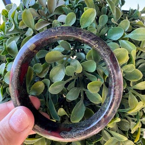 Peut inclure: Un bracelet jonc circulaire poli présentant un aspect sombre et tacheté avec des inclusions brun foncé, noires et rouge-marron subtiles sur toute la surface lisse et arrondie du matériau en pierre, créant un motif naturel et varié.