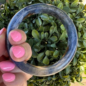 May include: A smooth, circular bangle bracelet crafted from translucent, dark blue and grey stone. The material features natural, cloudy marbling throughout its polished surface, creating a semi-transparent appearance with subtle variations in color depth.
