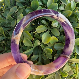 Könnte beinhalten: Ein polierter, kreisförmiger Armreif aus durchscheinendem violettem Amethyststein. Der Armreif weist natürliche Farbvariationen auf, darunter tiefviolette Töne, hellere lavendelfarbene Abschnitte und undurchsichtige weiße Mineraleinschlüsse im gesamten Band.