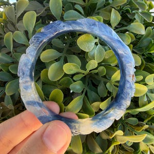 May include: A circular bangle bracelet crafted from translucent blue and white stone, featuring a carved, textured surface with undulating ridges and smooth, polished sections that create a marbled appearance throughout the entire piece.