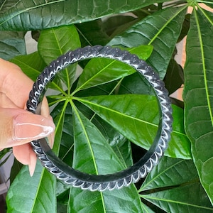 May include: A circular black bangle bracelet featuring a continuous twisted rope-like texture along its entire outer surface. The bracelet has a smooth, polished finish that reflects light, creating subtle highlights along the ridges of the twisted design.