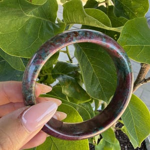 May include: A polished, round bangle bracelet with a marbled appearance. The bracelet features a mix of burgundy, teal, and brown tones. The jewellery is held in front of a backdrop of green leaves.