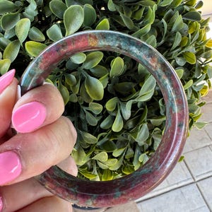 Puede incluir: Una pulsera tipo brazalete circular y lisa hecha de piedra pulida, con un patrón moteado de tonos rojo carmesí intenso y verde bosque en todo el material, mostrando una apariencia natural y variada con un acabado brillante y reflectante.