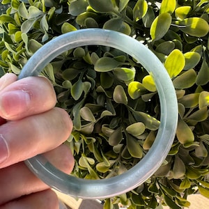 May include: A smooth, circular bangle bracelet crafted from translucent, pale green stone. The bangle features a polished, rounded surface with subtle variations in color, ranging from light mint to soft greyish-green tones throughout the material.