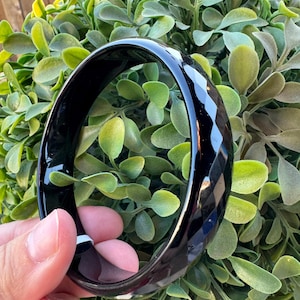 May include: A glossy black bangle bracelet featuring a faceted geometric surface design. The circular accessory has a smooth, reflective finish and a thick, solid band construction that creates a distinct contrast against the surrounding greenery.