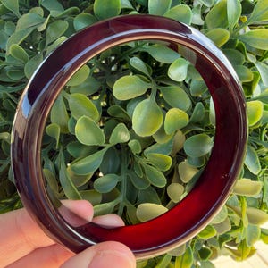 Puede incluir: Una pulsera tipo brazalete circular y lisa, fabricada con un material rojo oscuro translúcido. El brazalete presenta un acabado pulido y brillante que capta la luz, creando un brillo vibrante en toda la estructura sólida y continua del anillo.