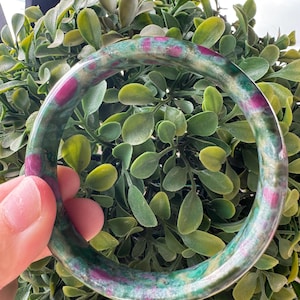 Può includere: Un bracciale rigido circolare realizzato in pietra lucida, caratterizzato da un motivo maculato con sfumature di verde bosco intenso e inclusioni rosa magenta vivaci su tutta la superficie liscia e arrotondata.