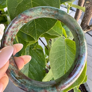 May include: A smooth, circular bangle bracelet crafted from polished stone, featuring a mottled pattern of deep forest green, teal, and earthy reddish-brown hues throughout the entire surface of the rounded, solid band.
