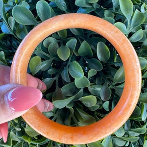 May include: A smooth, circular bangle bracelet in a solid, translucent orange hue. The bangle features a polished, rounded surface with a consistent thickness throughout its entire circumference, showcasing a uniform and vibrant warm orange color.