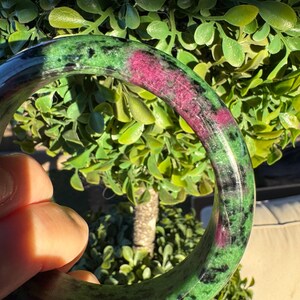 May include: A polished round bangle bracelet crafted from zoisite stone, featuring a vibrant mix of deep forest green, speckled black inclusions, and prominent patches of bright magenta-pink ruby throughout the smooth, circular surface.
