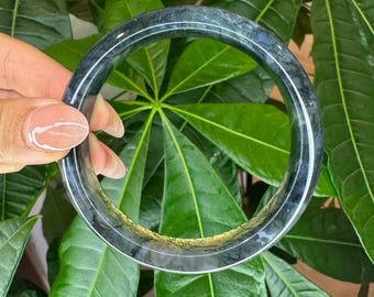 Może przedstawiać: Okrągła bransoletka typu bangle wykonana z ciemnego, nakrapianego zielonego kamienia o polerowanej, gładkiej powierzchni. Bransoletka posiada wyraźną, cienką, jasną linię biegnącą poziomo wokół środka, ukazującą naturalne wariacje minerałów.