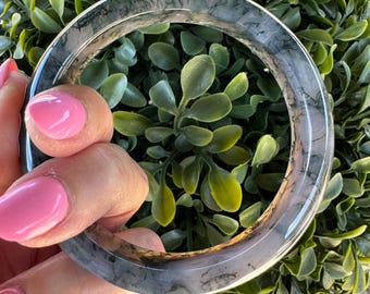Może przedstawiać: Okrągła bransoletka typu bangle wykonana z półprzezroczystego, czystego materiału z ciemnymi, dendrytycznymi, przypominającymi drzewa inkluzjami w całej strukturze. Bransoletka ma gładką, polerowaną powierzchnię i zaokrąglony profil.