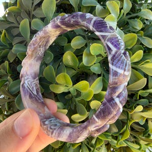 May include: A carved amethyst bangle bracelet featuring a twisted, rope-like texture with alternating bands of deep purple and opaque white mineral inclusions throughout the polished stone surface, creating a continuous circular design.