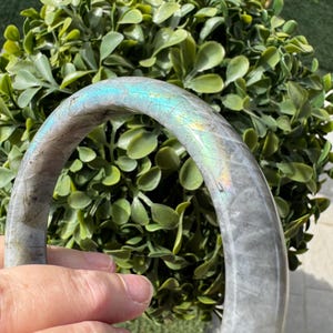 May include: A circular bangle bracelet crafted from polished labradorite stone, featuring a smooth, rounded surface with a grey base color and iridescent flashes of blue, green, and yellow light reflecting across the curved gemstone material.