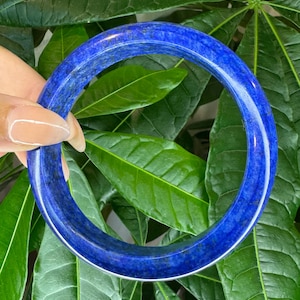 Puede incluir: Un brazalete circular tipo esclava fabricado en piedra lapislázuli pulida. La pulsera presenta un color azul profundo y vibrante con inclusiones oscuras naturales y sutiles motas doradas distribuidas por toda la superficie lisa y redondeada.