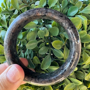 May include: A smooth, polished circular bangle bracelet crafted from a dark, translucent stone featuring mottled black and grey inclusions throughout its rounded surface, showcasing a natural, glossy finish with varying depths of color.