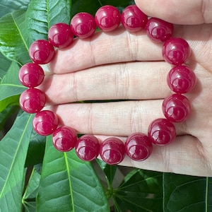 May include: A circular bracelet composed of smooth, polished, deep magenta-red spherical beads. The beads are uniform in size and strung together in a continuous loop, showcasing a glossy, reflective surface texture throughout the entire piece.