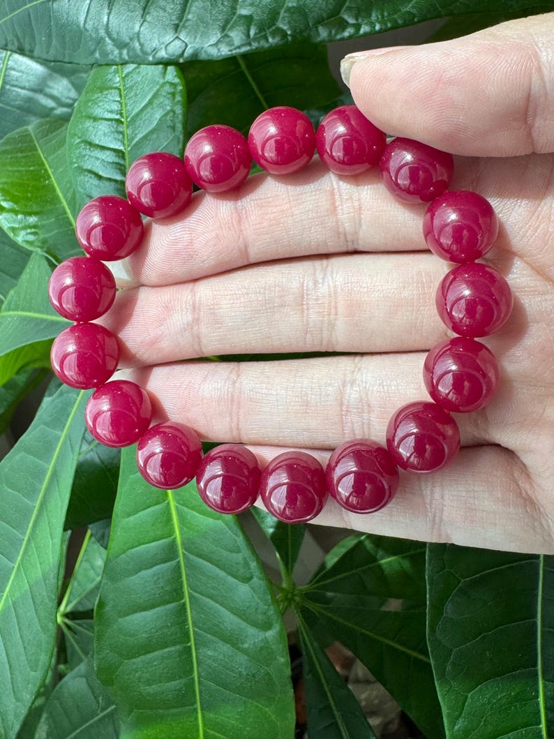 May include: A circular bracelet composed of smooth, polished, deep magenta-red spherical beads. The beads are uniform in size and strung together in a continuous loop, showcasing a glossy, reflective surface texture throughout the entire piece.