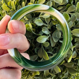 May include: A smooth, circular bangle bracelet featuring a translucent, polished finish with swirling patterns of deep forest green, lighter sage green, and creamy white tones throughout the entire rounded band structure.