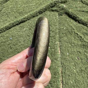 May include: A smooth, polished bangle bracelet crafted from golden obsidian. The stone features a reflective, metallic sheen with a deep black base color and a shimmering, bronze-gold luster that catches the light across its rounded, curved surface.