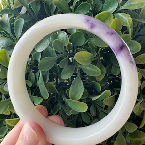 May include: A smooth, circular bangle bracelet crafted from polished white stone, featuring a distinct, natural patch of deep purple coloration along one side of the band. The bangle has a rounded profile and a glossy, reflective surface finish.