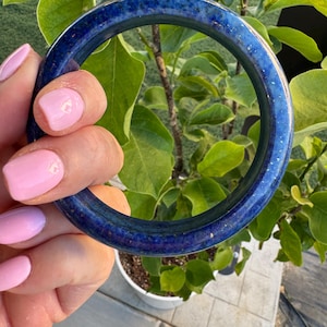 May include: A circular bangle bracelet featuring a deep blue color with visible gold-toned flecks throughout the surface. The bracelet has a smooth, polished finish and a rounded profile, showcasing a consistent thickness around the entire circumference.