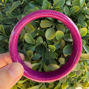 May include: A circular bangle bracelet featuring a deep magenta and purple gradient finish with a smooth, polished surface. The bracelet has a rounded profile and a consistent thickness throughout its entire circumference, showcasing a vibrant colour.