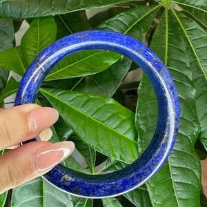 May include: A circular bangle bracelet crafted from deep blue lapis lazuli stone, featuring natural gold-colored pyrite flecks and dark mineral inclusions throughout the polished, smooth surface of the solid, continuous gemstone band.
