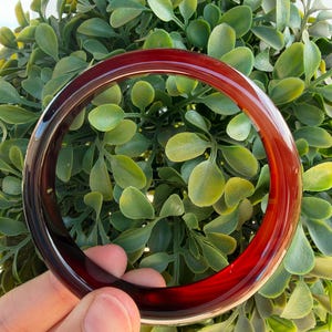 Może przedstawiać: Gładka, okrągła bransoletka typu bangle wykonana z półprzezroczystego, głęboko czerwonego materiału. Bransoletka ma polerowane, błyszczące wykończenie, które odbija światło, ukazując bogaty, ciemnokrwisty odcień przechodzący w jaśniejszą czerwień.