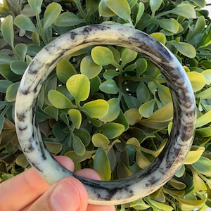 May include: A circular bangle bracelet featuring a smooth, polished surface with a marbled pattern of creamy off-white and deep charcoal black streaks. The accessory has a rounded profile and a glossy finish, showcasing natural-looking colour variations.
