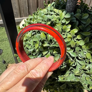 May include: A circular bangle bracelet featuring a glossy, vibrant red finish with a thin, dark charcoal-colored line running horizontally through the center of the band, creating a distinct contrast against the bright red surface.