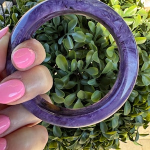 Puede incluir: Una pulsera tipo brazalete circular y lisa de color morado intenso con patrones jaspeados y arremolinados en todo el material. El brazalete tiene un acabado pulido y brillante con un perfil grueso y redondeado que forma un aro sólido.