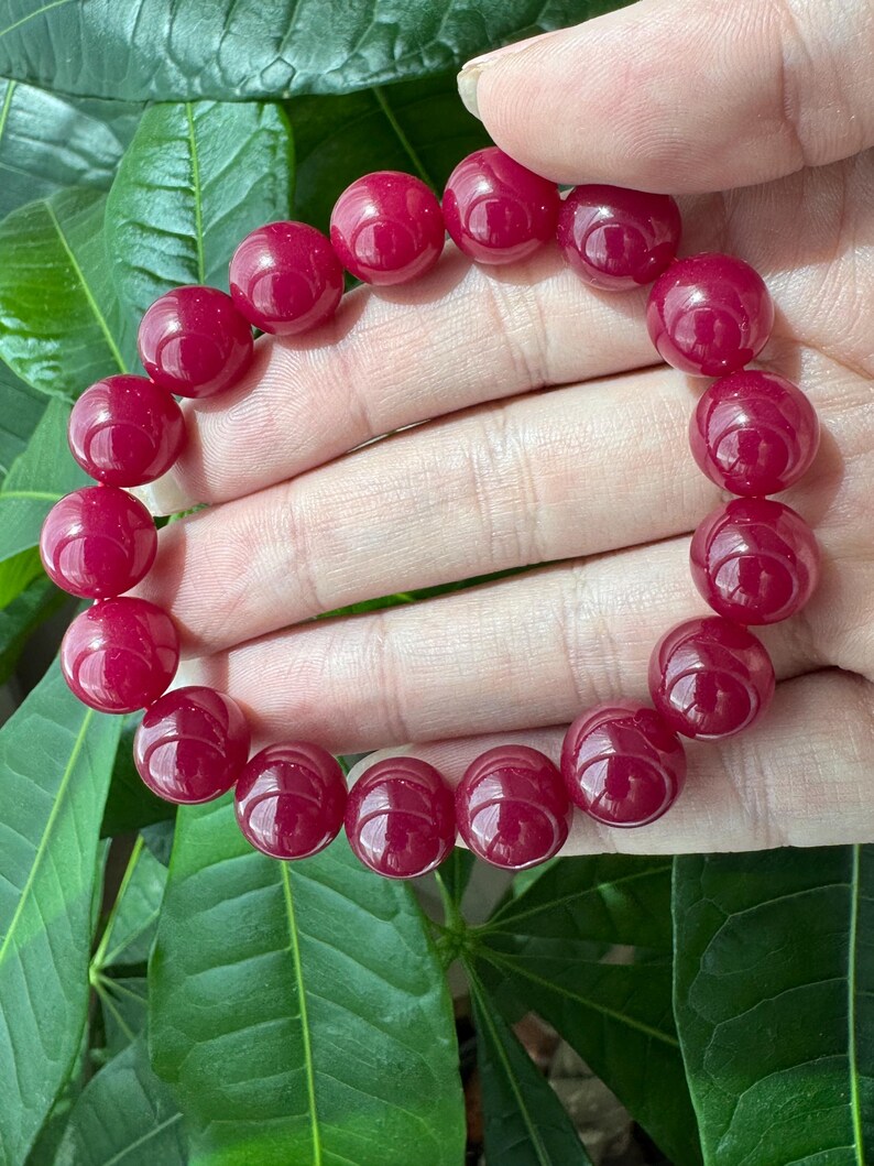 May include: A stretch bracelet composed of numerous smooth, round, deep magenta-colored beads. The beads are uniform in size and shape, featuring a polished, glossy finish that reflects light across their spherical surfaces in a circular arrangement.