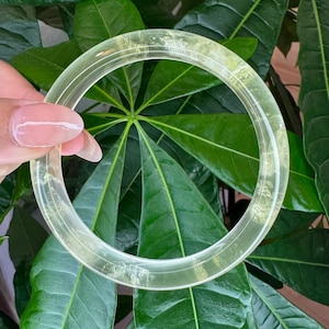 May include: A translucent, pale yellow bangle bracelet with a smooth, polished surface. The circular accessory features subtle internal inclusions and a clear, glass-like appearance, showcasing a consistent thickness throughout its rounded frame.