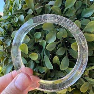 Peut inclure: Un bracelet jonc circulaire et transparent. Ce bracelet présente une surface lisse et polie avec de subtiles inclusions internes qui captent la lumière, créant un aspect cristallin et réfléchissant sur l'ensemble de l'anneau.