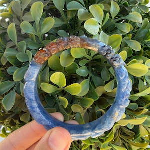 May include: A circular bangle bracelet featuring a textured blue surface with carved details. The upper section of the bangle is embedded with small, translucent amber-colored stones, while the rest of the band displays a matte blue finish.