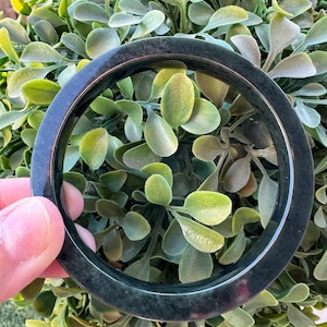 May include: A smooth, circular bangle bracelet with a dark, mottled black and deep green appearance. The surface of the bangle has a polished, reflective finish that catches the light, showcasing subtle variations in its dark, solid color tone.