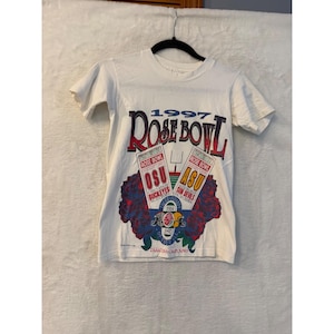 May include: A white short-sleeve t-shirt featuring a 1997 Rose Bowl graphic. The design includes two ticket stubs for OSU Buckeyes and ASU Sun Devils, red roses, a football goalpost, and the text Pasadena California printed at the bottom.