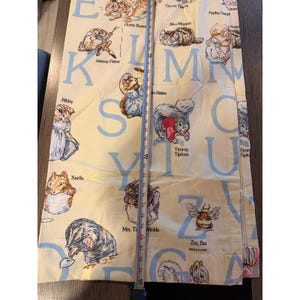 May include: A pale yellow fabric featuring a repeating alphabet pattern with illustrations of Beatrix Potter characters like Jeremy Fisher, Ribby, and Timmy Tiptoes. A metal measuring tape is placed vertically, showing a length of 60 inches.