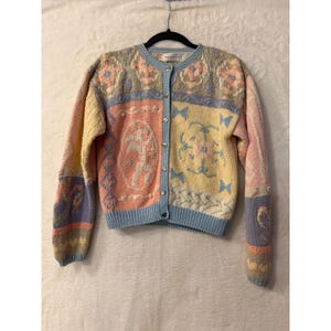 May include: A long-sleeved knit cardigan featuring a pastel colour palette of pink, yellow, and light blue. The garment displays intricate patterns including a central cherub motif, floral designs, and heart-shaped buttons along the front closure.