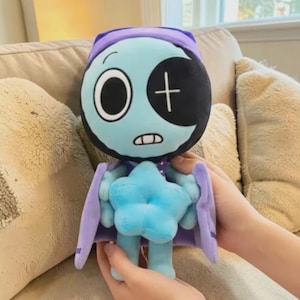 May include: A plush doll with a light blue face, a black eye patch, and a white cross. The doll has a purple cap and cape, and a blue star-shaped body. The doll is being held by a person.