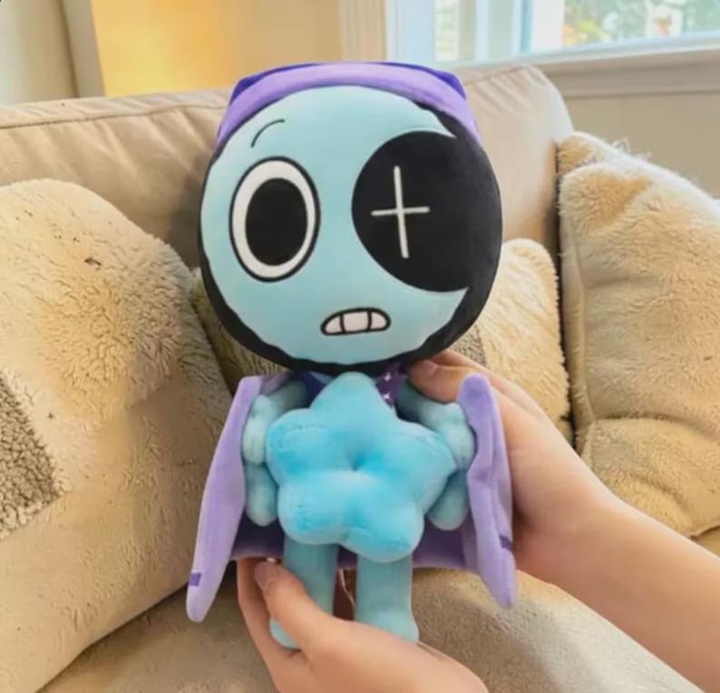 May include: A plush doll with a light blue face, a black eye patch, and a white cross. The doll has a purple cap and cape, and a blue star-shaped body. The doll is being held by a person.