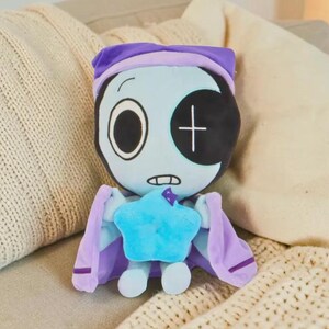 May include: A plush doll with a light blue face, one large open eye, and a black patch over the other eye. It wears a purple nightcap and cape, and holds a blue star. The doll is designed to look like a character.