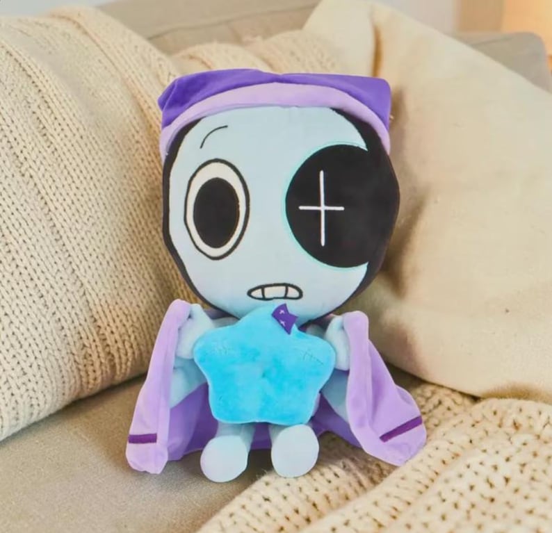 May include: A plush doll with a light blue face, one large open eye, and a black patch over the other eye. It wears a purple nightcap and cape, and holds a blue star. The doll is designed to look like a character.