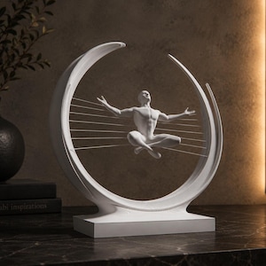 May include: A white sculpture featuring a central figure in a seated pose with arms outstretched, suspended within a curved, crescent-shaped frame. Thin horizontal lines connect the figure to the frame, creating a balanced, symmetrical composition.