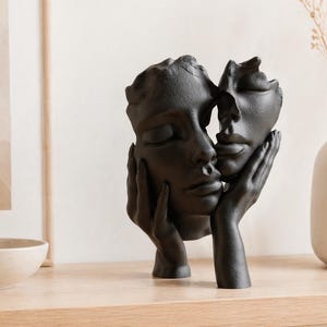 May include: A black sculpture of two faces, one resting against the other, with closed eyes. The sculpture is held by a pair of hands. The artwork is displayed on a wooden surface, with a vase and framed artwork in the background.