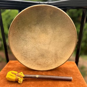 May include: A circular frame drum with a tan, textured animal hide head. Below the drum lies a mallet featuring a dark brown wooden handle and a bright yellow fabric-covered head secured with a thin leather cord wrapped around the base.