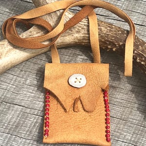 May include: A small, tan leather pouch with a long strap, adorned with a large white button and red beads along one side. The pouch is rectangular with a flap closure. The strap is made of the same tan leather. The background is a weathered wooden surface.
