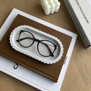 May include: A pair of round-framed eyeglasses with a dark brown frame and gold-colored arms rests on a white, scalloped-edge tray. The tray sits atop a stack of books, including a brown book with the word "CA" visible.