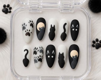 May include: A set of ten press-on nails with a cat theme. Designs include paw prints, cat silhouettes, and a cat with a moon. The nails are oval-shaped and come in black and white colors, packaged in a clear plastic case.